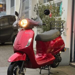 Rode scooter met hoog windscherm en brandende koplamp, geparkeerd voor een werkplaats.