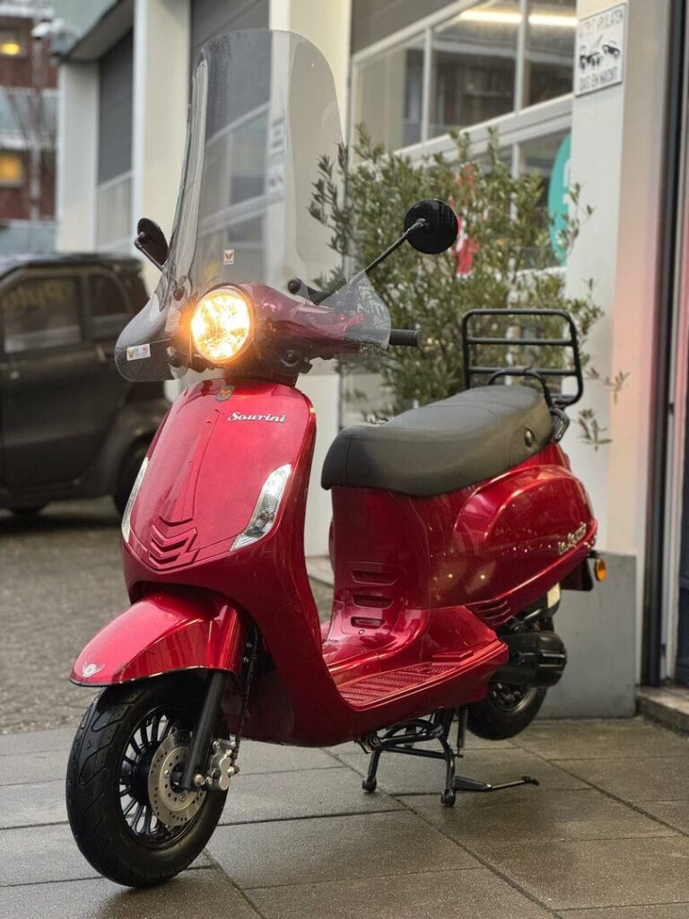 Rode scooter met hoog windscherm en brandende koplamp, geparkeerd voor een werkplaats.