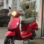 Rode scooter met windscherm en brandende koplamp, geparkeerd voor een werkplaats