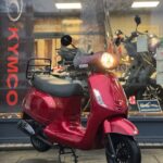 Rode retro-scooter met windscherm voor etalage van Kymco-dealer, koplamp brandt.