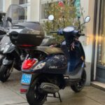 Donkerblauwe Fiddle III scooter met SHAD topkoffer, geparkeerd voor een winkel op een natte stoep.