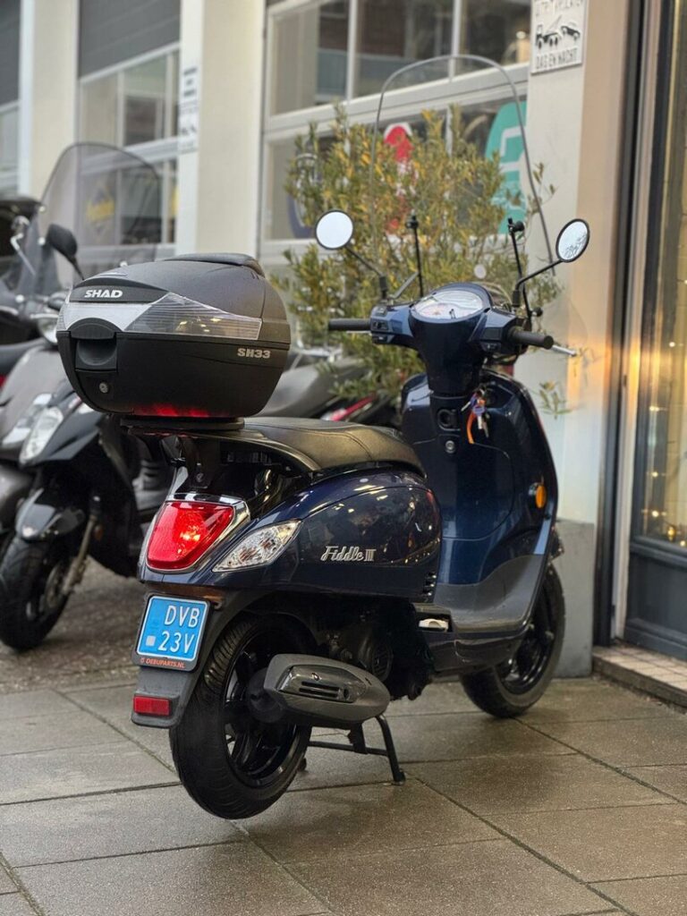 Donkerblauwe Fiddle III scooter met SHAD topkoffer, geparkeerd voor een winkel op een natte stoep.