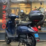 Donkerblauwe scooter met windscherm en topkoffer voor een motorshowroom geparkeerd.