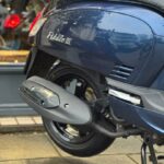 Close-up van de achterkant van een blauwe Fiddle III scooter met uitlaat en achterwiel op natte stoep.