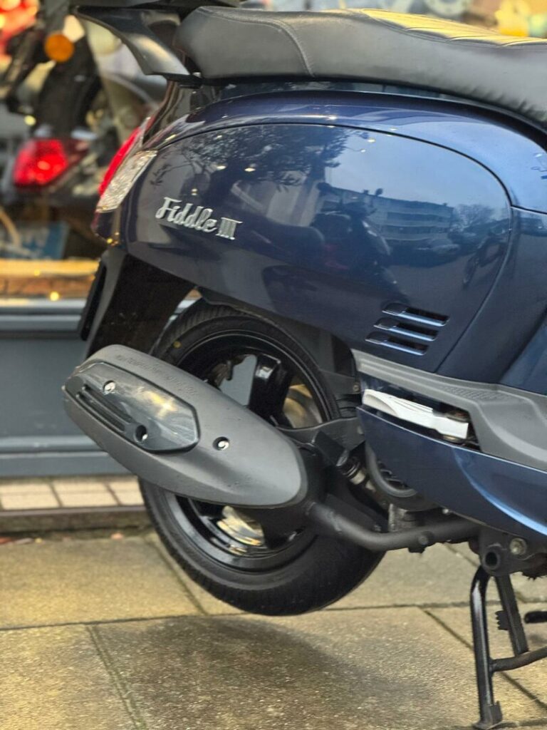 Close-up van de achterkant van een blauwe Fiddle III scooter met uitlaat en achterwiel op natte stoep.