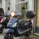 Donkerblauwe scooter met windscherm en topkoffer, geparkeerd voor een winkel.