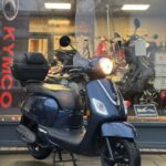Donkerblauwe scooter met windscherm en topkoffer voor een motorshowroom.