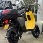 Gele en grijze elektrische scooter met topkoffer, geparkeerd op de stoep bij een winkel.