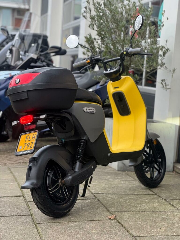 Gele en grijze elektrische scooter met topkoffer, geparkeerd op de stoep bij een winkel.