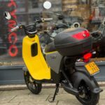 Gele scooter met topkoffer geparkeerd voor een winkelraam met KYMCO-tekst.