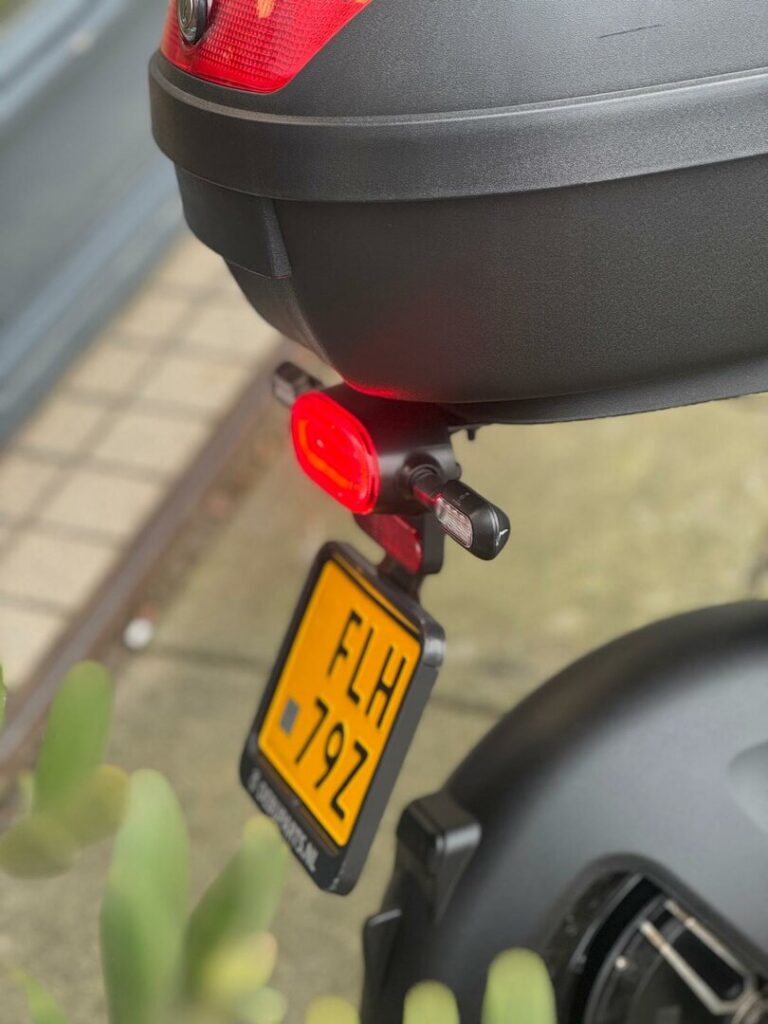 Achterkant van een scooter met topkoffer, brandend achterlicht en gele kentekenplaat.