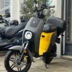 Zwart-gele elektrische scooter met ronde koplamp geparkeerd op stoep voor winkel