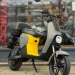 Gele en grijze elektrische scooter met topkoffer, geparkeerd op de stoep voor een winkelraam.