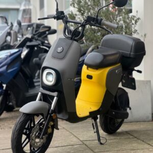 Zwart-grijze elektrische scooter met gele treeplank en topkoffer, geparkeerd op de stoep.