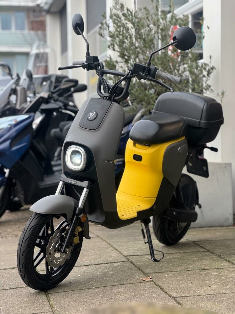 Zwart-grijze elektrische scooter met gele treeplank en topkoffer, geparkeerd op de stoep.