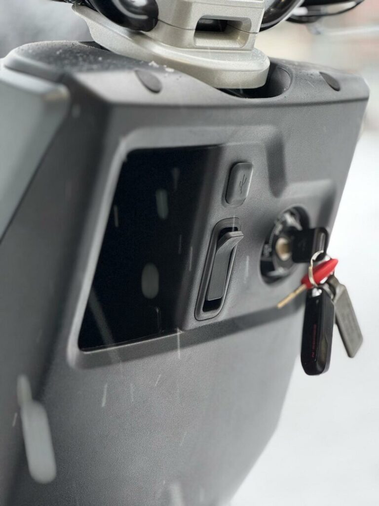 Close-up van scooterdashboard met contactslot, sleutelbos, USB-poort en opbergvak.