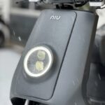 Close-up van de voorzijde van een grijze NIU e-scooter met ronde LED-koplamp en regendruppels.