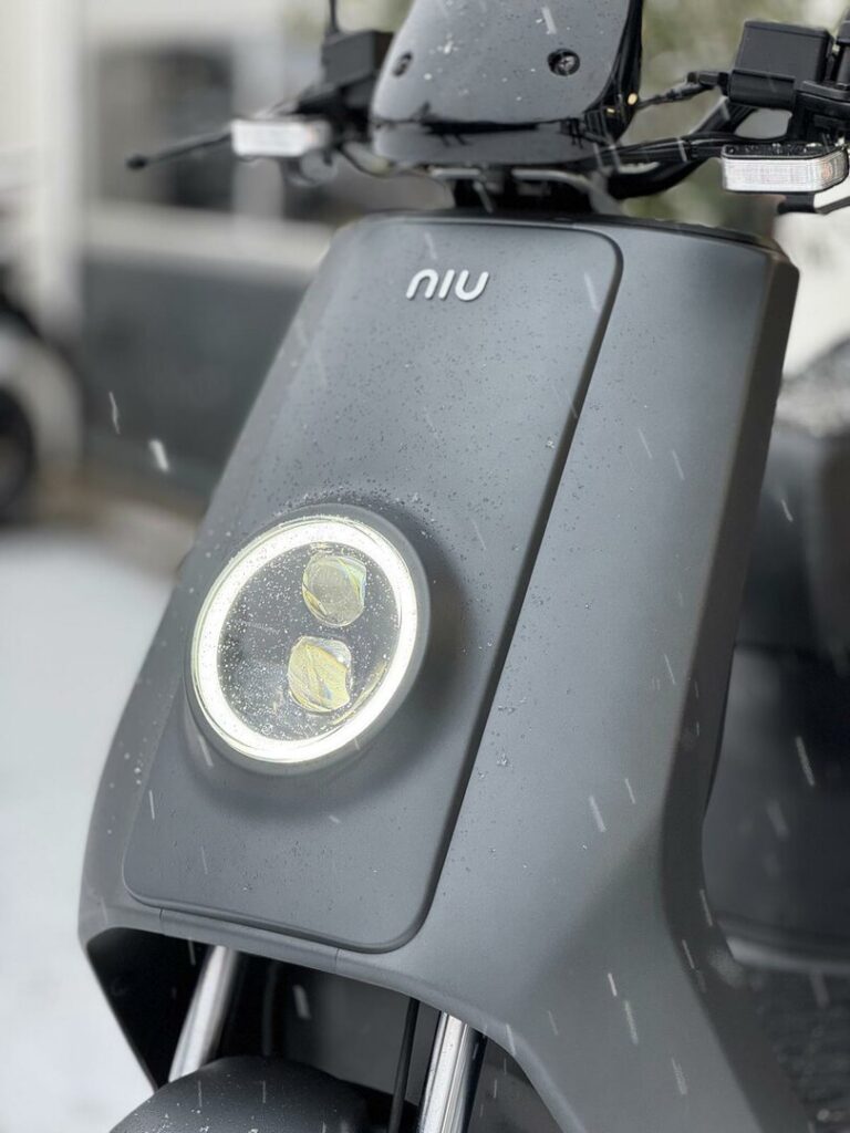 Close-up van de voorzijde van een grijze NIU e-scooter met ronde LED-koplamp en regendruppels.