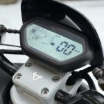 Close-up van het digitale motorfietsdashboard met 0 km/u en 28% batterij, met druppels op de stuurklem.