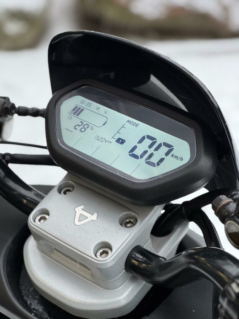 Close-up van het digitale motorfietsdashboard met 0 km/u en 28% batterij, met druppels op de stuurklem.