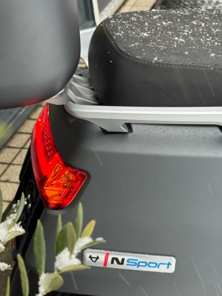 Close-up van de achterkant van een grijze scooter met N Sport-logo, brandend achterlicht en sneeuw op het zadel.