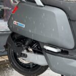 Achterwiel en zijkant van een grijze scooter in de sneeuw, met vallende sneeuwvlokken en zichtbare schijfrem.