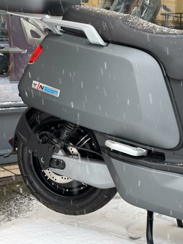 Achterwiel en zijkant van een grijze scooter in de sneeuw, met vallende sneeuwvlokken en zichtbare schijfrem.