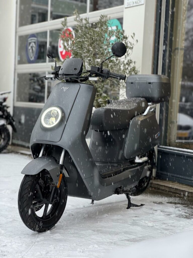 Grijze elektrische NIU-scooter met topkoffer geparkeerd buiten in de sneeuw