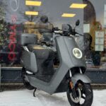 Grijze elektrische NIU scooter in de sneeuw voor een showroom met verlichte ramen.