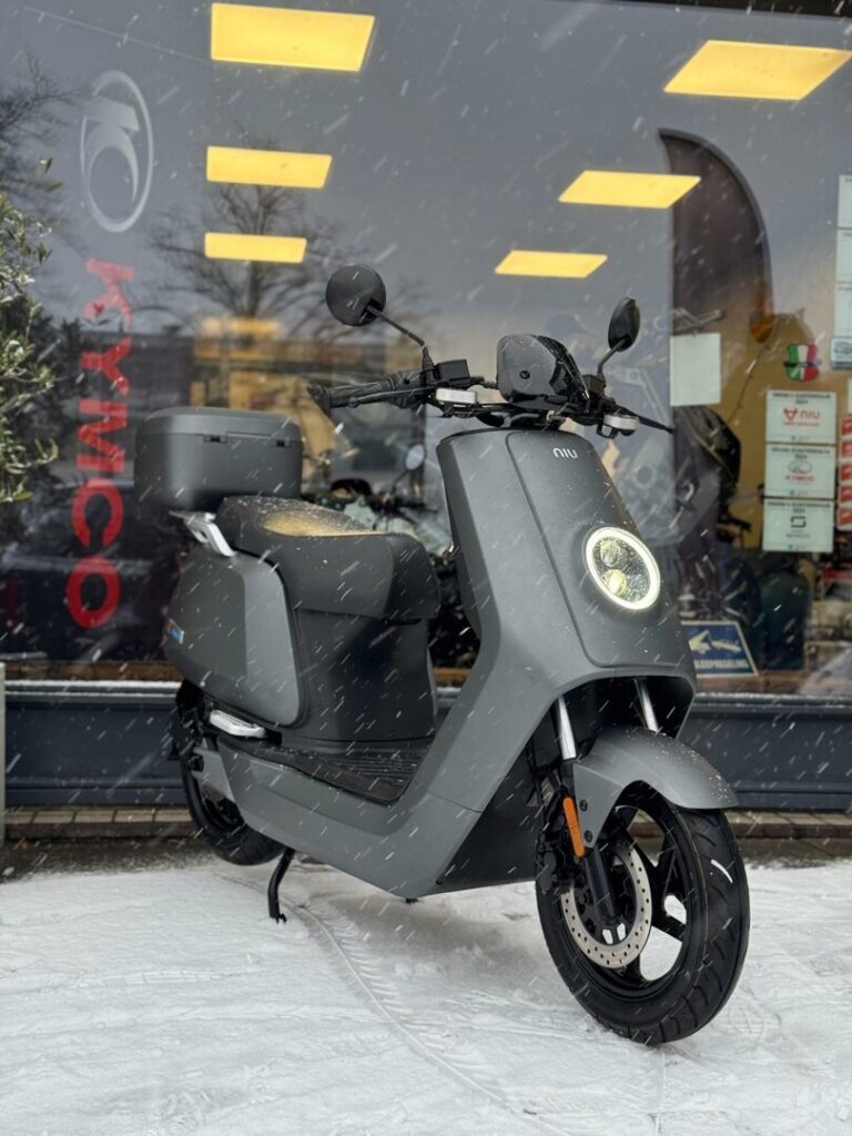 Grijze elektrische NIU scooter in de sneeuw voor een showroom met verlichte ramen.
