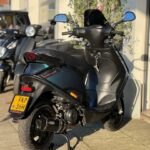 Zwarte scooter met gele kentekenplaat geparkeerd voor een winkel in de zon.