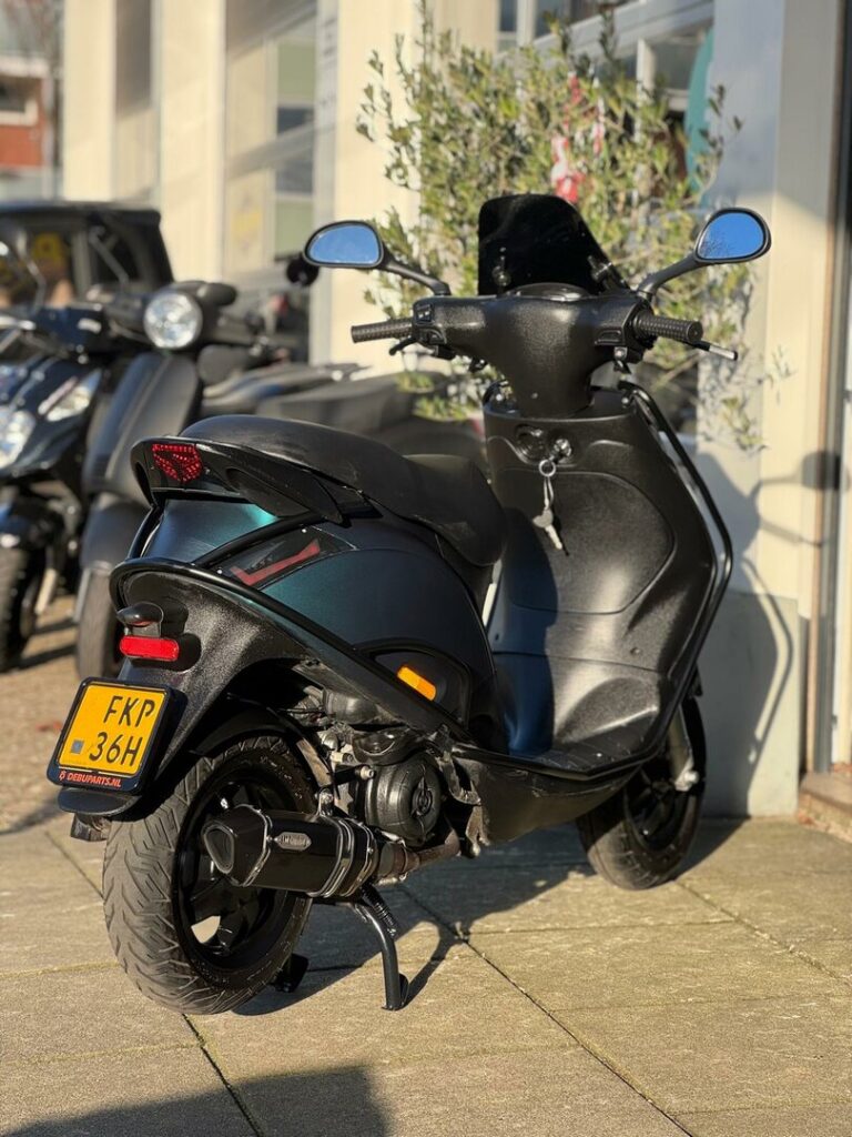 Zwarte scooter met gele kentekenplaat geparkeerd voor een winkel in de zon.