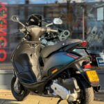 Zwarte motorscooter op standaard, geparkeerd op stoep voor een winkelruit op een zonnige dag.