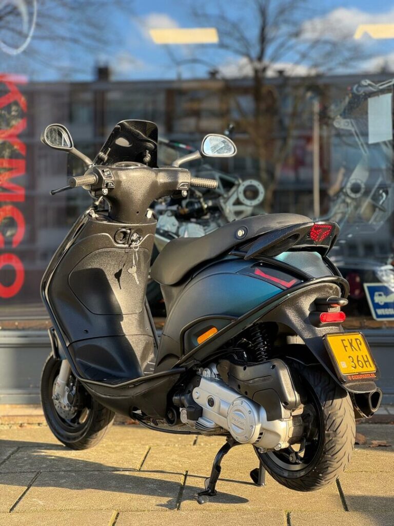 Zwarte motorscooter op standaard, geparkeerd op stoep voor een winkelruit op een zonnige dag.