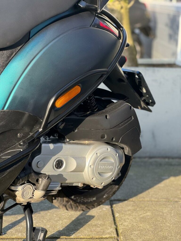 Close-up van de achterzijde van een scooter met Piaggio-motor, veer en achterwiel in warm zonlicht.