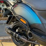 Close-up van de achterzijde van een blauwe scooter met JMTuning uitlaat, op de standaard geparkeerd.