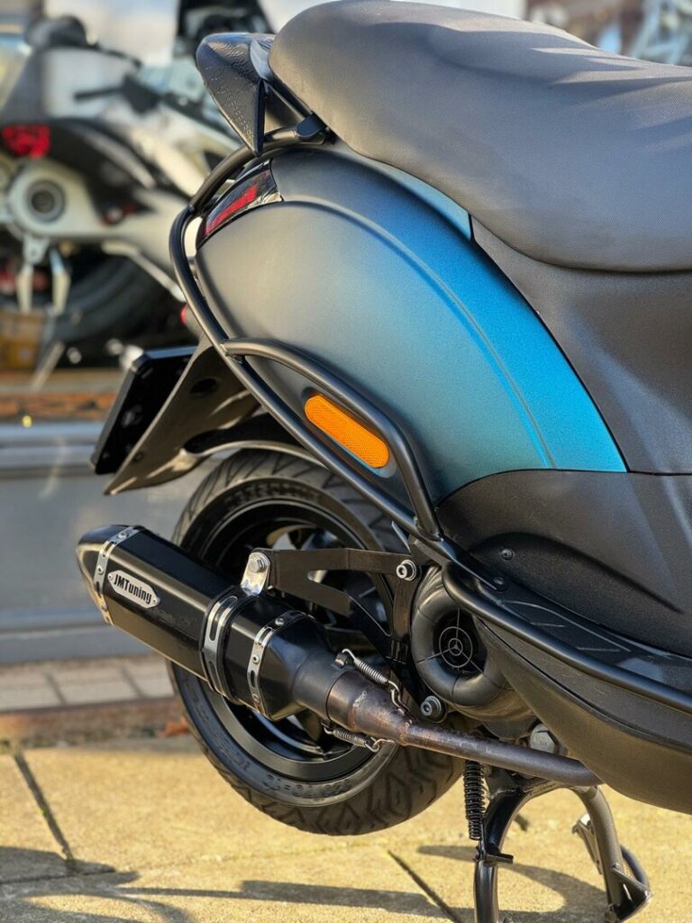 Close-up van de achterzijde van een blauwe scooter met JMTuning uitlaat, op de standaard geparkeerd.