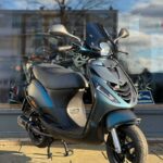 Matte zwarte scooter op middenbok voor winkelruit, in zonlicht met weerspiegelde lucht.