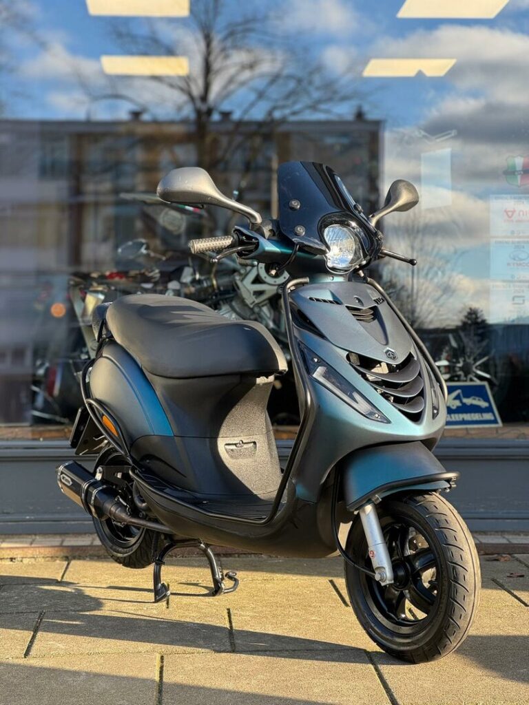 Matte zwarte scooter op middenbok voor winkelruit, in zonlicht met weerspiegelde lucht.