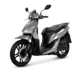 Zilverkleurige moderne scooter met hoge wielen en schijfrem, gefotografeerd schuin van voren op witte achtergrond.