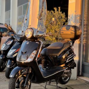 Zwarte scooter met hoog windscherm en topkoffer, geparkeerd voor een winkel in avondzon