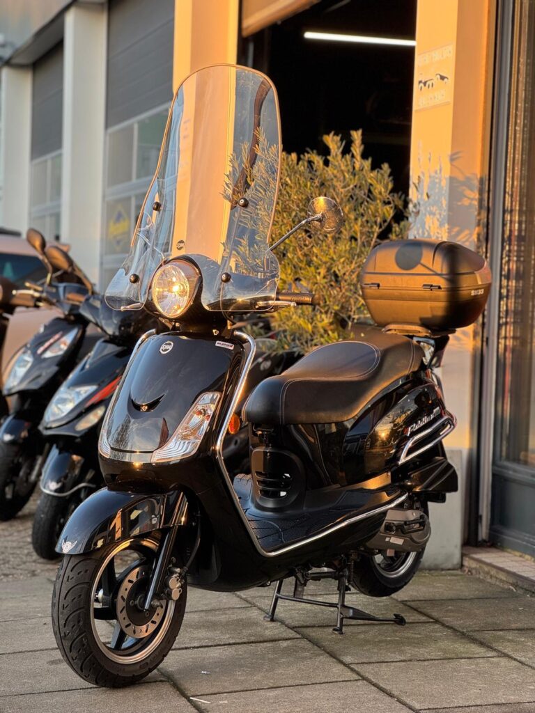 Zwarte scooter met hoog windscherm en topkoffer, geparkeerd voor een winkel in avondzon