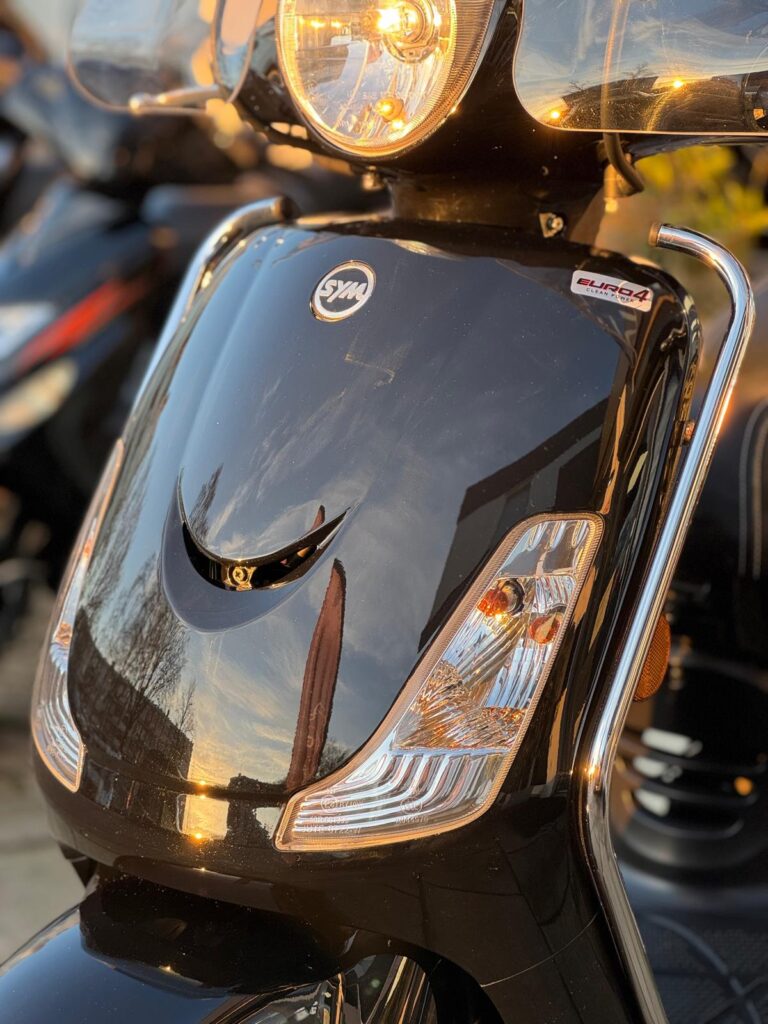 Close-up van de voorkant van een zwarte scooter met koplamp en knipperlichten in avondzon.