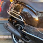 Close-up van de achterzijde van een glanzend zwarte scooter met zadel, uitlaat en achterwiel.