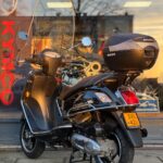 Zwarte scooter met windscherm en topkoffer, geparkeerd voor een winkelraam.