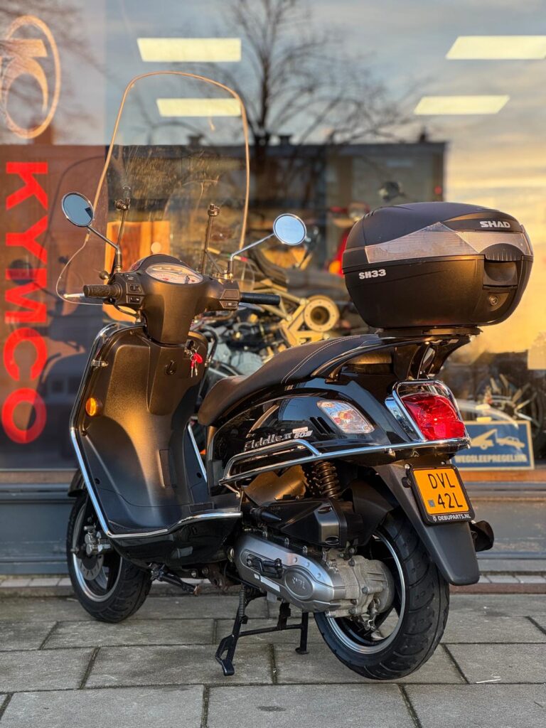 Zwarte scooter met windscherm en topkoffer, geparkeerd voor een winkelraam.