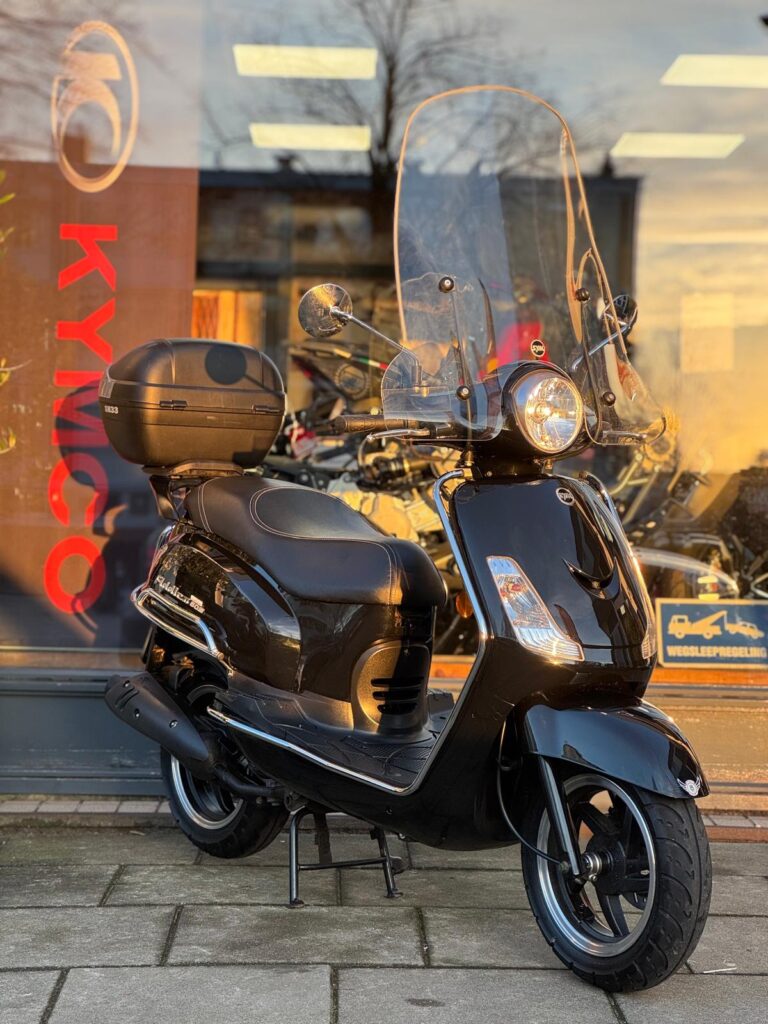 Zwarte scooter met hoog windscherm en topkoffer, op standaard voor een winkel met KYMCO-logo.
