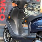 Close-up van een blauwe scooter op de standaard, bij een showroom met KYMCO-logo op de ruit.