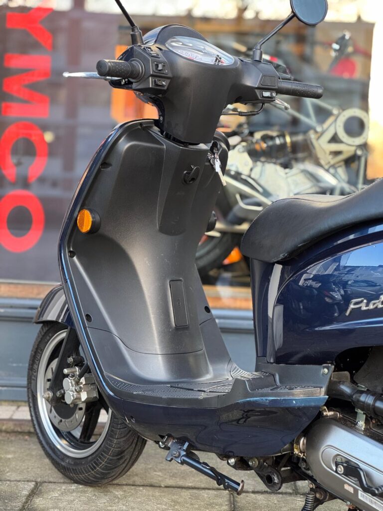 Close-up van een blauwe scooter op de standaard, bij een showroom met KYMCO-logo op de ruit.