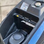 Open bergruimte onder het zadel van een blauwe scooter met onderhoudssticker en brandstofdop.